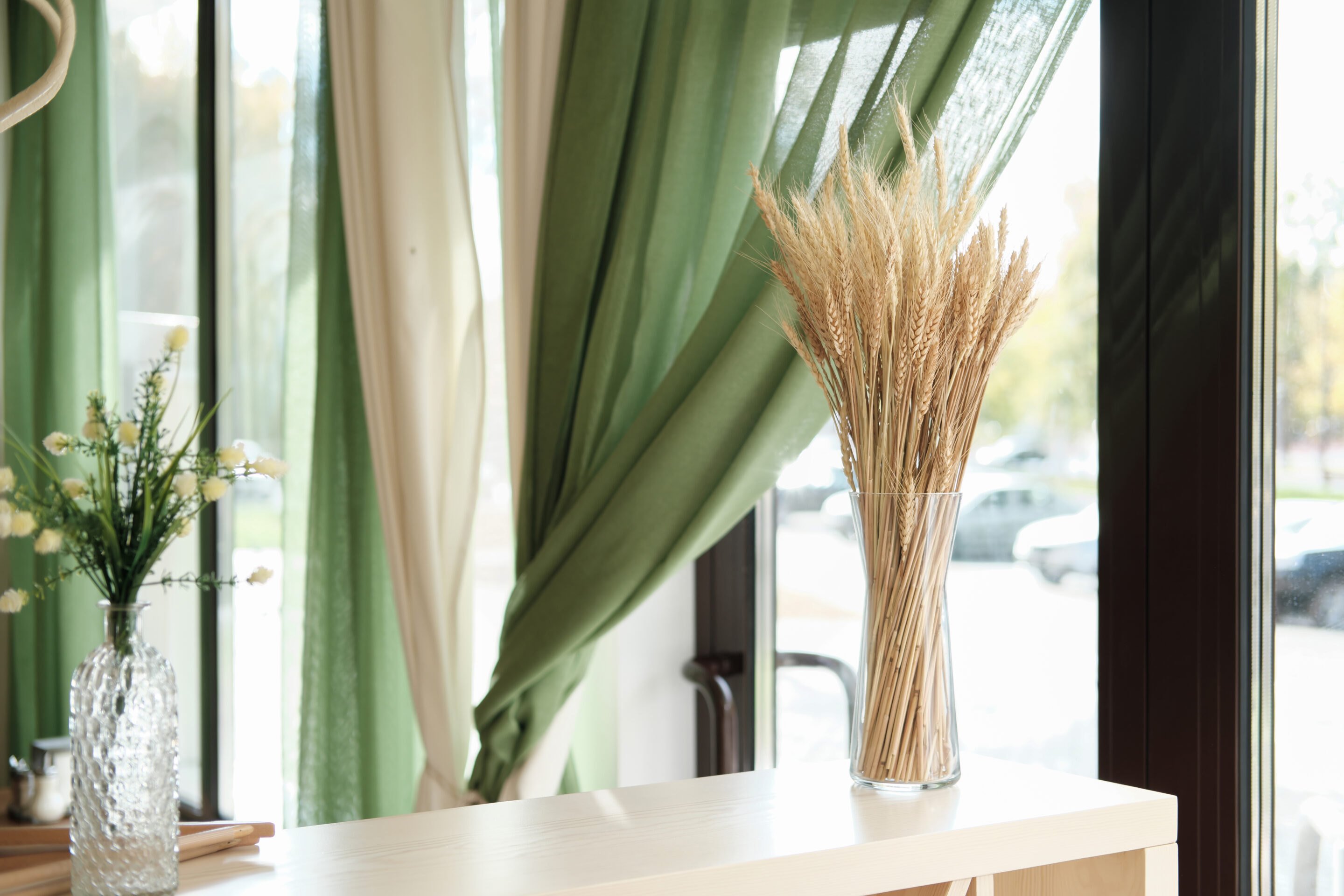 partial view of stylish interior of cafe or terrace with natural decor. ear of wheat in a glass vase on light wooden table, green curtains and flowers. window view to a street.